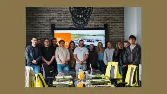 Grupa uczestników wizyty studyjnej stoi w sali konferencyjnej firmy. W tle widoczny jest ekran prezentacyjny z fotografią centrum logistycznego, a na pierwszym planie znajduje się stół konferencyjny z materiałami i kamizelkami odblaskowymi.