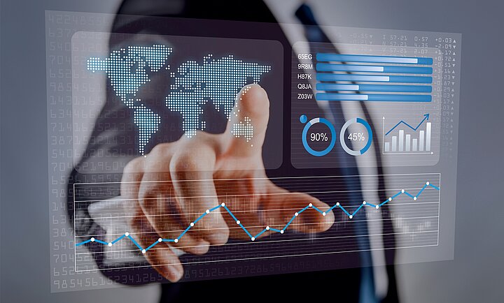 Osoba w garniturze wskazuje na przezroczysty interfejs cyfrowy z wizualizacjami danych: mapą świata, wykresami słupkowymi, kołowymi (90% i 45%) oraz wykresem liniowym z niebieskimi punktami.