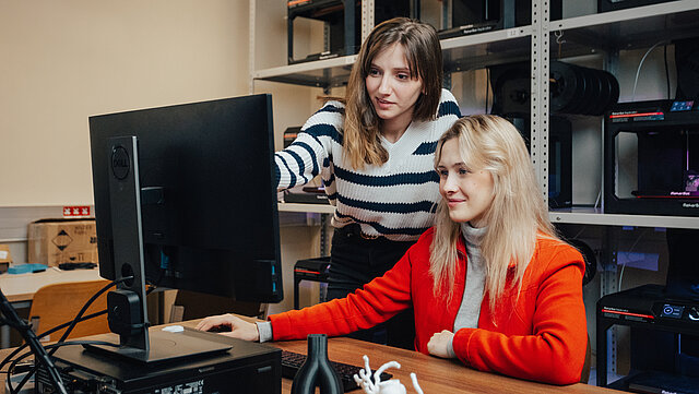 Dwie studentki obserwują z uwagą monitor komputera. Jedna wskazuje palcem cos drugiej