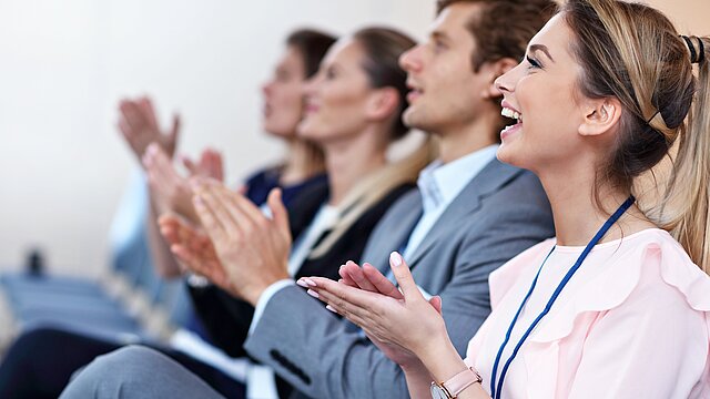 Grupa osób w strojach biznesowych siedzi w rzędzie i bije brawo. Scena sugeruje udział w konferencji lub seminarium.