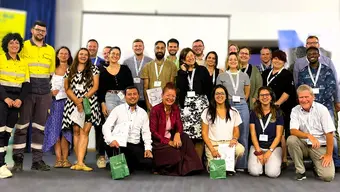 Zdjęcie grupowe uczestników konferencji lub wydarzenia. Osoby stoją i klęczą w rzędach, mają przypięte identyfikatory, niektórzy trzymają torby. W tle widoczny jest ekran projekcyjny, niebieskie zasłony oraz baner z częściowo nieczytelnym tekstem.