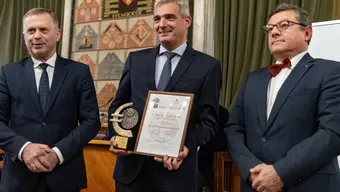 rzech mężczyzn w garniturach stoi obok siebie. Mężczyzna w środku trzyma oprawiony certyfikat oraz trofeum. W tle widoczna jest dekoracyjna tkanina z różnorodnymi wzorami i symbolami.