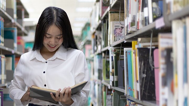 Zdjęcie studentki czytającej między półkami bibliotecznymi