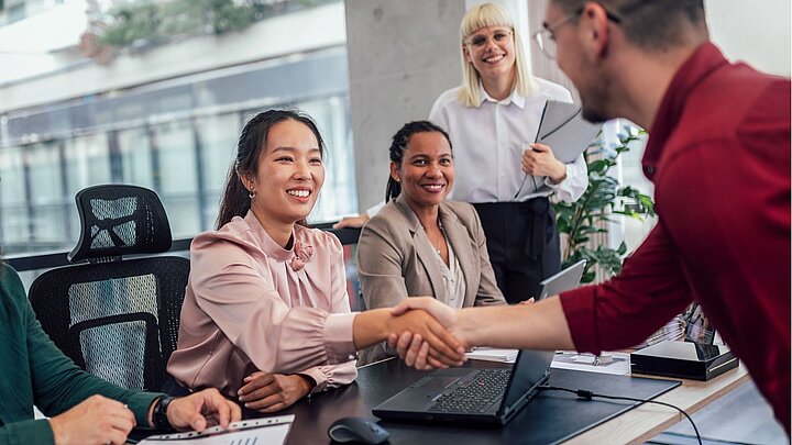 Zdjęcie grupy ludzi zgromadzonych przy stole spośród których dwie osoby witają się podając dłonie