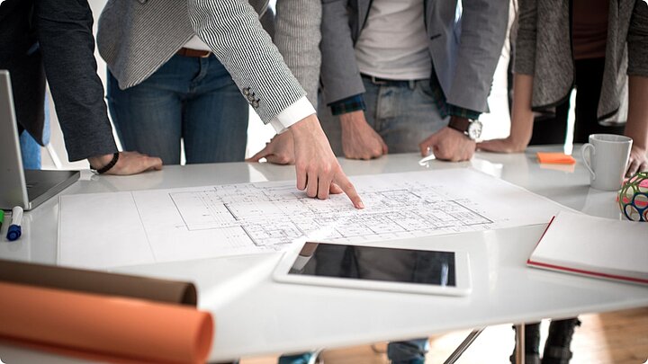 Zdjęcie grupy osób stojących przy stole z rozłożonymi dokumentami w trakcie dyskusji