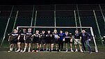 Grupa jedenastu osób stoi przed bramką piłkarską na oświetlonym boisku nocą. Większość osób ubrana jest w czarne stroje sportowe i ma na szyi medale. W centrum grupy widoczny jest dyplom, a po prawej stronie jedna z osób trzyma puchar. W tle znajduje się wysoka siatka otaczająca boisko oraz ciemne nocne niebo.