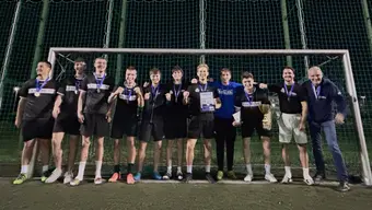 Grupa jedenastu osób stoi przed bramką piłkarską na oświetlonym boisku nocą. Większość osób ubrana jest w czarne stroje sportowe i ma na szyi medale. W centrum grupy widoczny jest dyplom, a po prawej stronie jedna z osób trzyma puchar. W tle znajduje się wysoka siatka otaczająca boisko oraz ciemne nocne niebo.