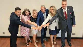 Grupa sześciu osób stoi w półokręgu, wyciągając ręce do środka i dotykając się nawzajem dłońmi. Tło jest jednolite, a podłoga drewniana.