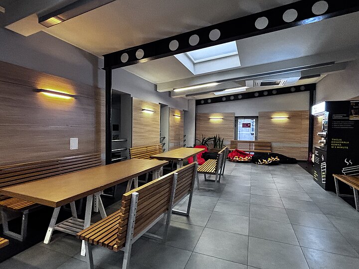 Przestrzeń z długimi stołami i ławkami ustawionymi w rzędach, oświetlona lampami ściennymi; w głębi widoczne pufy oraz rośliny.