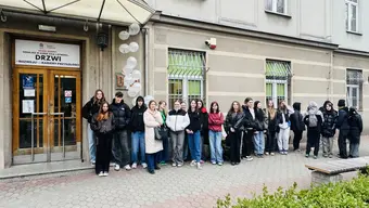 Grupa osób stoi w szeregu przed budynkiem z napisem nad wejściem: „DRZWI ROZWOJU – KLUCZEM PRZYSZŁOŚCI”. Przy wejściu znajdują się balony. Budynek ma betonową elewację z poziomymi pasami i okna zabezpieczone kratami.