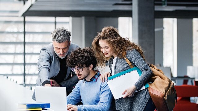 Zdjęcie dwóch studentów oraz nauczyciela przed komputerem
