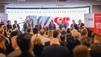Konferencja biznesowa z panelem prelegentów siedzących z przodu sali pełnej uczestników. W tle widoczny jest duży baner z napisem „STRATEGIC PARTNER IN BUSINESS EXCELLENCE” oraz „MASS”. Po bokach znajdują się dwa dodatkowe banery: po lewej „MINDSET LEADS”, po prawej „CRACOVIA”.