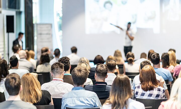 Grupa osób siedząca w sali konferencyjnej, zwrócona w stronę prelegenta stojącego z przodu. W tle widoczny rozmyty ekran projekcyjny, sugerujący trwającą prezentację
