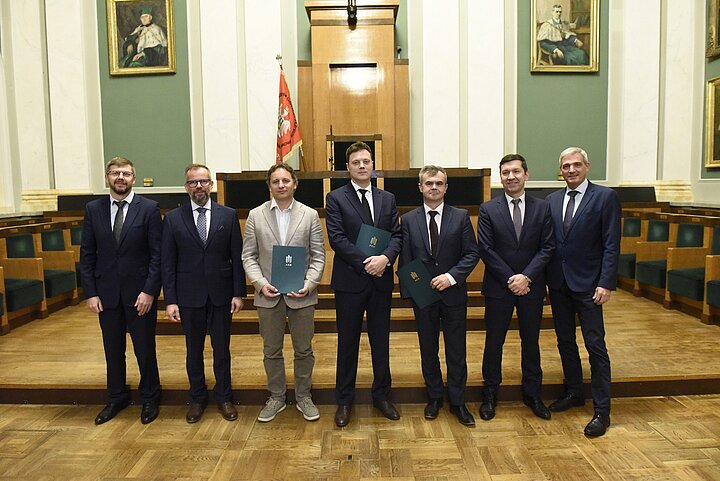 Siedmiu mężczyzn w formalnym otoczeniu, sześciu w garniturach, jeden w jasnej marynarce i szarych spodniach. Trzech z nich trzyma zielone teczki z godłem. W tle drewniane panele, zielone ściany, portrety i flaga z czerwonymi elementami