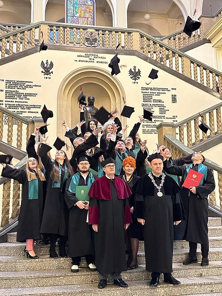 Grupa absolwentów świętuje, rzucając birety w powietrze na schodach wewnątrz budynku o ozdobnej architekturze.