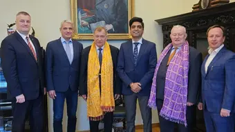 ześciu mężczyzn w formalnych strojach stoi w pomieszczeniu z drewnianą podłogą i ozdobnymi meblami. Dwóch z nich ma kolorowe szarfy — jedną żółtą, drugą fioletową. W tle widoczny jest duży portret starszego mężczyzny z siwymi włosami, trzymającego książkę.