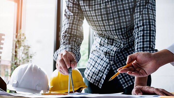 Zdjęcie inżyniera stojącego obok stołu na którym położone są dwa kaski budowlane