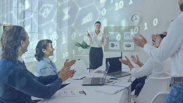 Osoba prowadząca prezentację w nowoczesnym biurze, stojąca przed grupą ludzi siedzących przy stole i bijących brawo. W tle widoczne są cyfrowe grafiki z ikonami (wykresy, klepsydra, chmura), kod binarny oraz zegar i wykresy na ekranie