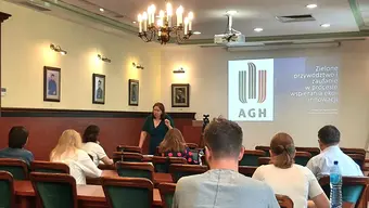 Sala wykładowa lub konferencyjna z uczestnikami siedzącymi i zwróconymi w stronę prelegenta stojącego przy mównicy. Na ekranie projektora widoczne jest logo AGH oraz tytuł prezentacji: „Zielone przywództwo i zarządzanie w procesie wspierania ekoinnowacji”.