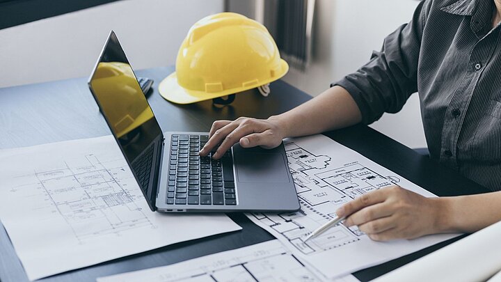 Zdjęcie dłoni mężczyzny na stole na którym znajdują się kask dokumenty i laptop