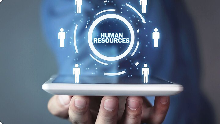 Zdhęcie dłoni podtrzymującej tablet nad którym wygenerowany jest cyfrowo napis hjuman resourses otoczony kołem z ikonami