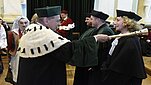 Osoba w uroczystym stroju akademickim z biało-czarnym futrzanym kołnierzem dotyka ceremonialnym berłem ramienia innej osoby ubranej w czarną togę i biret podczas akademickiej ceremonii. W tle widoczne są kolejne osoby w togach oraz uczestnicy w strojach ludowych we wnętrzu reprezentacyjnej sali.