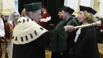 Osoba w uroczystym stroju akademickim z biało-czarnym futrzanym kołnierzem dotyka ceremonialnym berłem ramienia innej osoby ubranej w czarną togę i biret podczas akademickiej ceremonii. W tle widoczne są kolejne osoby w togach oraz uczestnicy w strojach ludowych we wnętrzu reprezentacyjnej sali.
