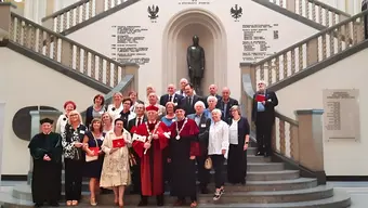 Grupa osób w strojach formalnych, w tym ceremonialnych togach i łańcuchach, stoi na schodach w budynku podczas oficjalnego wydarzenia. W tle widoczna jest statua umieszczona w niszy oraz tablice pamiątkowe z inskrypcjami i datami.