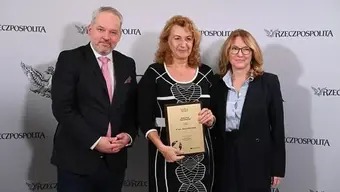 Trzy osoby stoją przed ścianką z napisem „RZECZPOSPOLITA” i godłem orła. Osoba w centrum trzyma tabliczkę lub certyfikat.