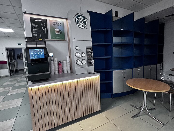Automat Starbucks ustawiony obok niebieskich regałów i małego stolika; w tle widok na korytarz i drzwi.